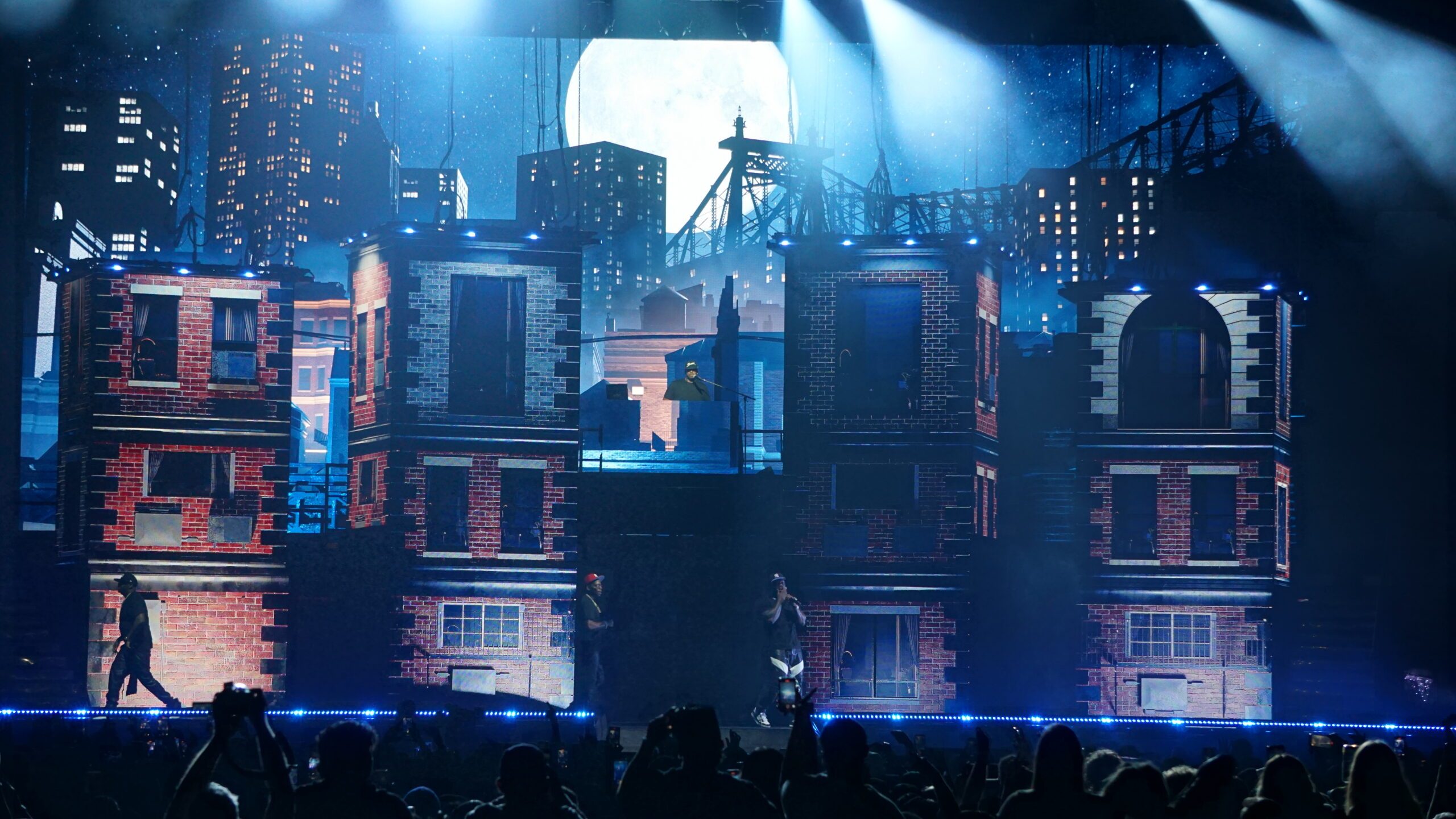 Brick buildings under a full moon and a New York bridge at night, with the band on stage
