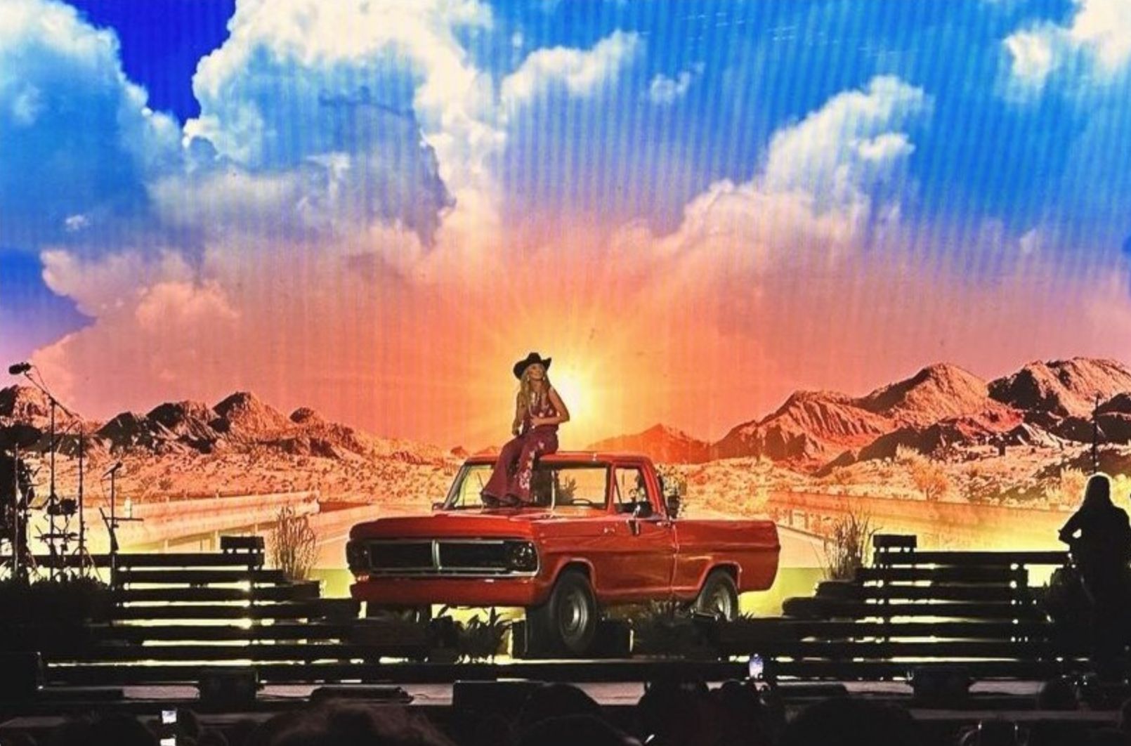 Lainey Wilson sitting on a red pickup truck in front of a desert sunset on the screens