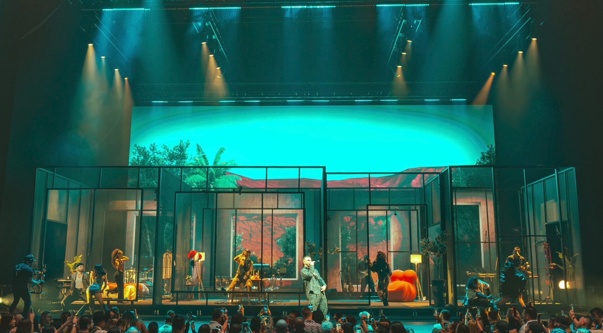 Wide view of the stage: a glass house set in front of a teal sky and red hills on the LED wall