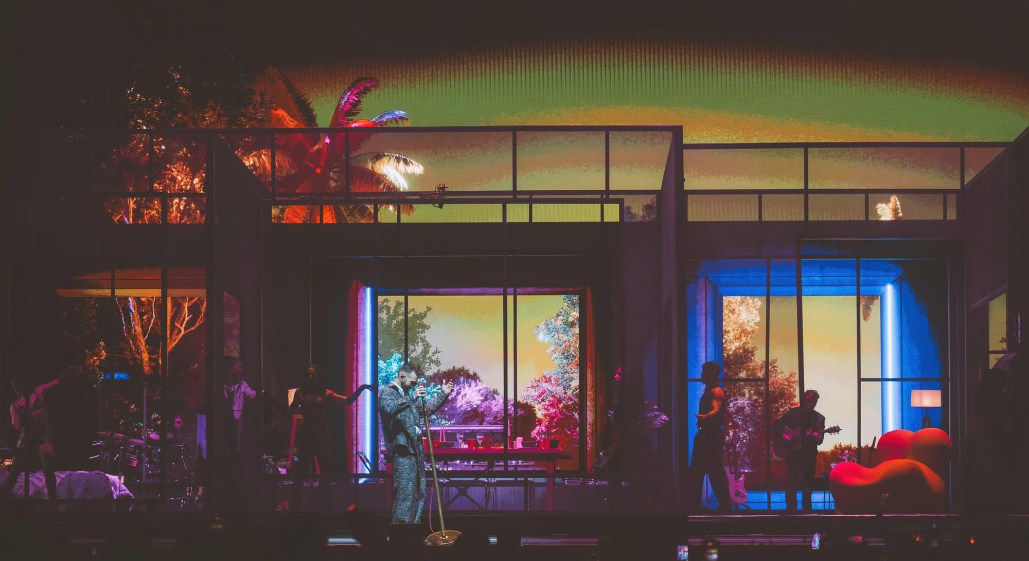 The glass house at night, its windows glowing with neon-colored palms and trees