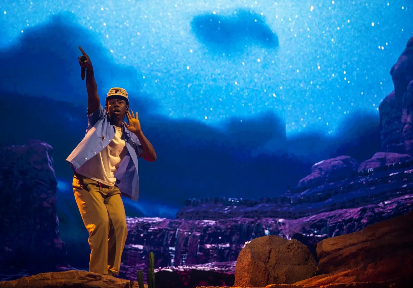 Tyler, the Creator pointing skyward in front of a starry blue desert canyon