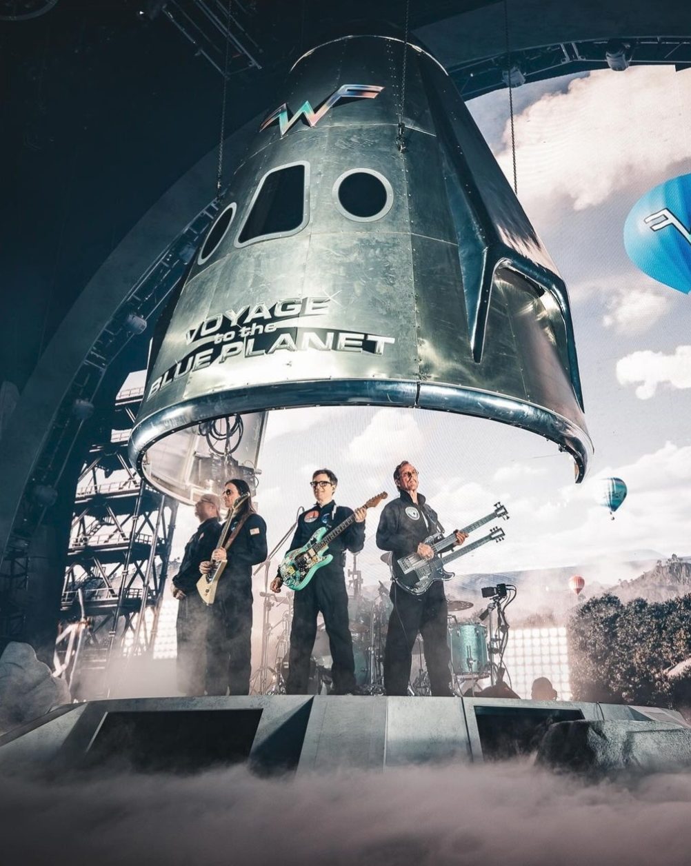 Weezer performing under a giant Voyage to the Blue Planet space capsule, with balloons on the screens behind